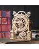 The 3D дървен пъзел Ugears - Старинен будилник (43 части) с видими зъбни колела и ажурни украси е поставен върху книгата, а на заден план се виждат размазани рафт с книги и камера.-6-thumb