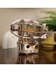 The 3D дървен пъзел Ugears - Стиймпънк Дирижабъл, 170 части с видими зъбни колела и ластици стои върху полиран плот, а на заден план се виждат зелено растение, бял скулптурен обект и жълта ваза.-3-thumb