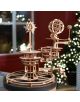 3D дървен пъзел Ugears - Tellurion Слънце, Земя, Луна, 249 части стои на маса на закрито, показвайки своите зъбни колела и спираловидна рампа. На заден план се виждат размазани празнични светлини и дърво.-6-thumb
