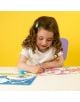 Младо момиче с къдрава коса и синя щипка за коса седи на бяла маса и щастливо оцветява Подложка за оцветяване Floss & Rock - Rainbow Fairy със син маркер, усмихнато и съсредоточено на яркожълт фон.-5-thumb