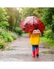 Дете в жълт дъждобран и червени ботуши върви по мокра пътека, държейки Детски чадър Legami - Ladybug с формата на усмихната божа краставица. Буйна зеленина ги заобикаля в този дъждовен ден.-6-thumb