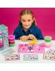 Младо русо момиче се наслаждава на своя Детски творчески комплект Floss & Rock с глина Rainbow Fairy, оформяйки еднорози и животни върху розова подложка. Наблизо са разположени ванички с глина и карти с инструкции, всичко това на розов фон.-5-thumb