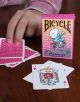Ръка държи карти за игра до кутия Карти за игра Bicycle Brosmind Four Gangs с карикатурен велосипедист. На дървената маса валето и дамата на сърцата разкриват живи, причудливи илюстрации от това уникално тесте.-4-thumb