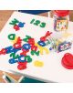 Пластмасовите букви и цифри от Комплект букви и цифри на Apli Kids са разпръснати на бяла маса до два отворени контейнера, а на заден план се виждат столовете и дървеният под на книжарница Orange.-2-thumb