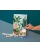 Една ръка рисува Комплект за рисуване по номера Graine Creative - Градска джунгла 20 x 30 см., изобразяваща растения в саксии. Малки саксии с боя и още две четки от комплекта лежат върху сивата повърхност до платното.-4-thumb