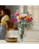 В прозрачна стъклена ваза е изложен букетът „LEGO Botanicals Красив букет с розови цветя“. До нея стои декоративна купа с капак, а на замъгления фон се вижда човек, стоящ до осветена от слънцето стена.-7-thumb