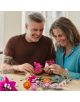 Усмихната двойка на средна възраст сглобява комплекта „LEGO Botanicals: Красив букет с розови цветя“ на масата си, заобиколена от готови цветя, в весела и творческа атмосфера.-5-thumb