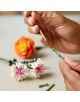 Две ръце сглобяват зелени LEGO елементи от комплекта „LEGO Botanicals: Красив букет с розови цветя“, за да образуват стъбло на цвете, като на заден план се виждат красив букет от розови цветя и други готови цветя от LEGO.-6-thumb