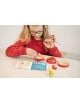 Малко дете с очила и червена риза си играе с логическа игра Smart Games: Пъзел в ябълка, поставяйки цветни фигурки вътре, докато проверява инструкциите на бяла маса.-3-thumb
