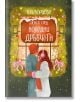 Двойка в зимни дрехи стои рамо до рамо до празнично украсена витрина, заснемайки истинската коледна романтика. Над тях "Любов сред коледни дръвчета" показва името на продукта на български език.-2-thumb