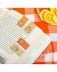 Отворена книга с три Kikkerland магнитни книгоразделители - Капибара на страниците си, поставена върху оранжево-бяла карирана кърпа до чиния с нарязани цитрусови плодове.-3-thumb