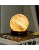 Настолната лампа Gingko AstraGlass Light Mini Polar Cloud с мраморен жълто-кафяв мотив и дървена основа грее топло, наподобявайки планета, поставена върху затворена книга с частично видим текст на корицата.-2-thumb