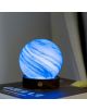 Светещ синьо-бял настолна лампа Gingko AstraGlass Light Mini Polar Cloud с вихрови шарки, изработен от ръчно издухано стъкло, се намира върху тъмна дървена основа върху затворена книга, в която се вижда част от заглавието EVYBRO.-3-thumb