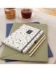 Седмичният тефтер-органайзер Miquelrius Grafica Terrazo Plus 2026 е поставен върху зелена и синя папка, до два молива, а на заден план има чаша чай и буркан с кафява захар върху бяла повърхност.-7-thumb