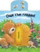 Симпатичен гладно мече седи до голям жълт кошер, протягайки ръка за мед, докато таралеж стои на зелена трева с цветя, а две пчели летят над него. Детската книжка "Още съм гладен" е на български език, със синьо небе и облаци.-thumb