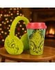 Зелена чаша за път Fizz Creations - The Grinch с червен капак седи на табуретка до размити зелените ушанки Fizz Creations - The Grinch, а размазани коледни лампички и камина създават празнична, уютна атмосфера.-3-thumb