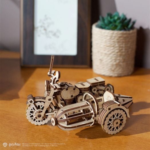 3D дървен пъзел Ugears Хари Потър - Летящият мотор на Хагрид, 130 части е изложен върху дървена повърхност с фина изработка, до рамкирана снимка и малко растение в оплетена саксия на заден план.-8