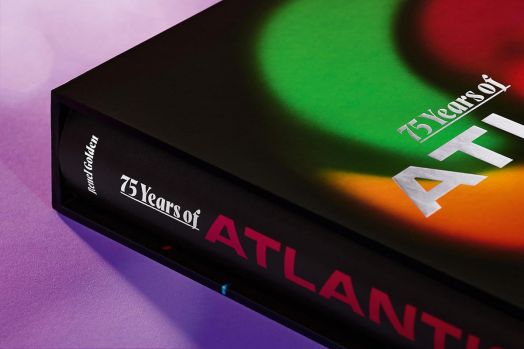 Близък план на книгата "75 Years of Atlantic Records", със смели, цветни надписи на корицата и гръбнака, лежи върху лилава повърхност и е частично поставена в черно калъфче.-2