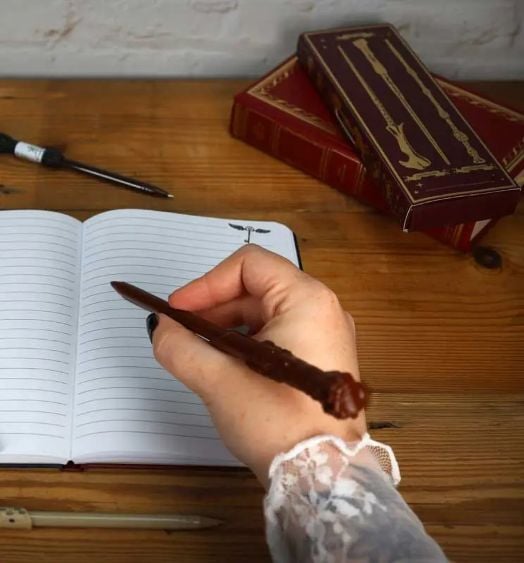 Една ръка държи Комплект химикалки магически пръчици Blue Sky Harry Potter, 4 бр. писалка над отворена тетрадка върху дървено бюро, а на заден план има две червени книги и друга писалка, които напомнят за магията на Хари Потър.-5