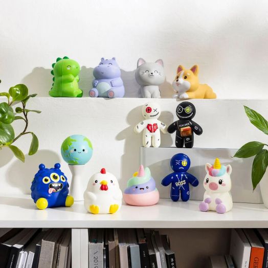 Колекция от цветни фигурки на играчки - животни, чудовища, еднорози - плюс Антистрес фигурка Legami - Corgi, изложени на бели рафтове със зелени растения до тях и книги, които се виждат частично на заден план.-6