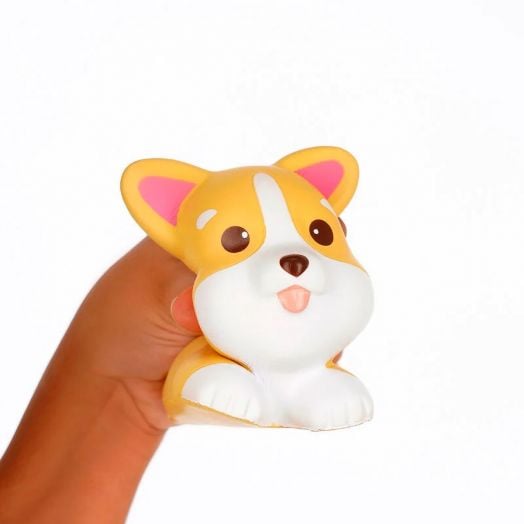 Една ръка стиска Антистрес фигурка Legami - Corgi, сладка жълто-бяла плюшена играчка с формата на корги с големи очи, розови уши и изплезен език, на обикновен бял фон.-2
