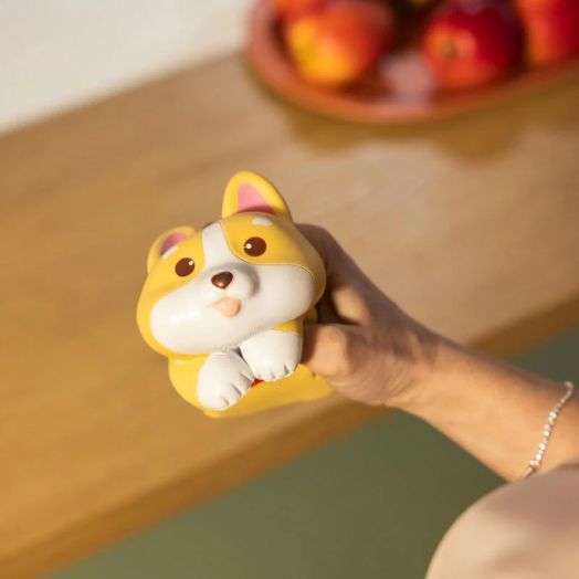 Човек държи Антистрес фигурка Legami - Corgi, сладка кръгла играчка с големи очи и малко езиче, а на заден план върху дървена повърхност почиват ябълки.-5