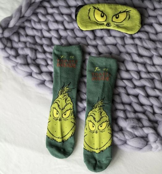 Чорапи и маска за сън Fizz Creations - The Grinch: комплект меки чорапи с лика на Гринч и надпис "Im 99% TOASTY INSIDE", плюс маска за сън с очи на Гринч, подредени върху лавандулено одеяло.-5