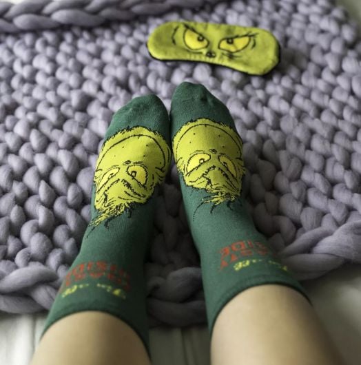 Човек в меки чорапи от Чорапи и маска за сън Fizz Creations - The Grinch с лицето на Гринч седи върху дебело лилаво одеяло, а маската за сън със зелените очи на Гринч почива наблизо.-6