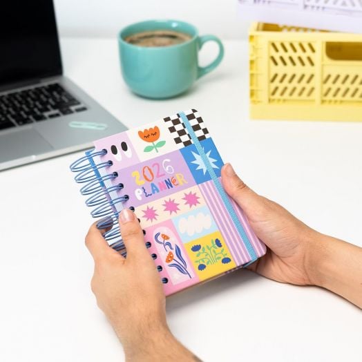 Човек държи Дневен тефтер-органайзер Grupo Erik Dopamine 2026 на бюро с лаптоп, зелена чаша за кафе и жълта щайга на заден план.-2