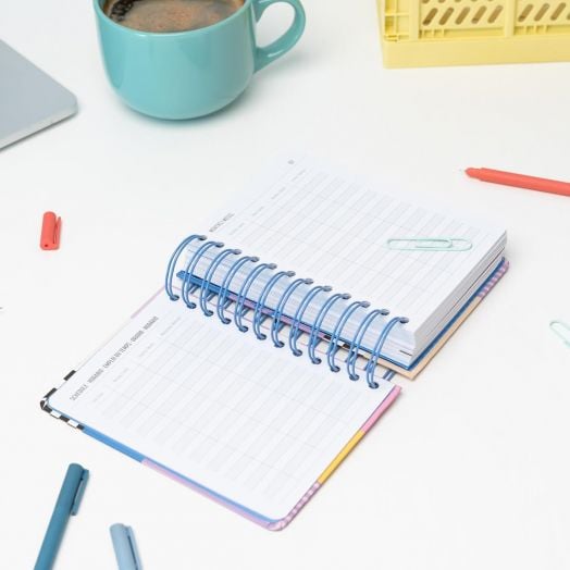 Отворен Дневен тефтер-органайзер Grupo Erik Dopamine 2026 лежи на бяло бюро, заобиколен от цветни химикалки, кламери, чаша в тиландски цвят, жълта кутия и част от лаптоп. Празните му страници очакват вашите планове за 2026 г.-4