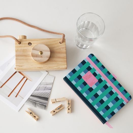 Дървен фотоапарат-играчка, чаша с вода, две отпечатани снимки, четири дървени щипки за дрехи и джобен седмичен тефтер - органайзер Miquelrius Grafica Color Block 2026 са подредени грижливо върху бяла повърхност.-6