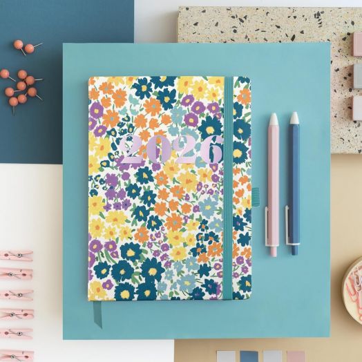 Джобният седмичен тефтер-органайзер Miquelrius Natura Impressionist 2026 с флорална корица е поставен върху син фон, обграден от два молива, щипки, кабърчета и пастелни геометрични листа.-6