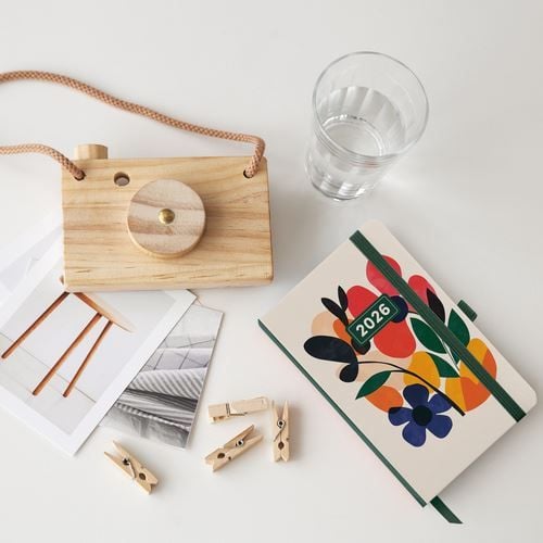Плоскост с дървена играчка фотоапарат, чаша с вода, Джобен тефтер - Оргайзер Miquelrius Natura Mini Garden 2026 с цветна флорална корица, отпечатани снимки и дървени щипки върху бяла повърхност.-3