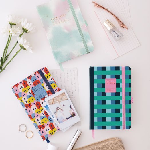 Плоска подредба на четири цветни дневника, включително Джобен тефтер - Оргайзер Miquelrius Grafica Color Block 2026, бели маргаритки, златна писалка, прозрачен лак за нокти, две моментни снимки и ръкописна бележка върху бяла повърхност.-7