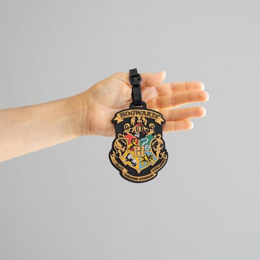Ръката държи Етикет за багаж Grupo Erik Harry Potter - Hogwarts с герба на Хогуортс с четирите домашни животни и девиза, което прави този Етикет за куфар задължителен аксесоар за всеки фен на Хари Потър.-7