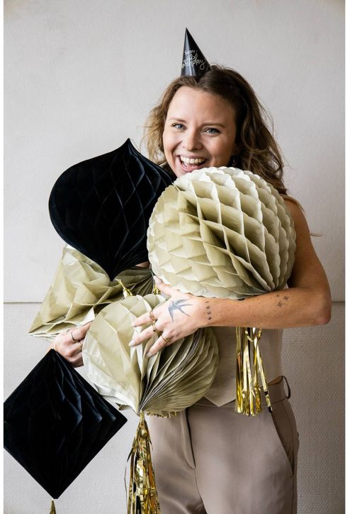 Усмихната жена с шапка "Честит рожден ден" държи в ръцете си големи медени топки и пискюли от Хартиена декорация Folat - Crème Noir, 3 бр., застанали на фона на обикновена стена - идеална парти декорация за всеки украса за рожден ден.-4