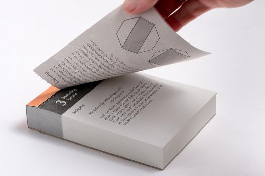 Една ръка прелиства дебелата книжка "Heureka Mathematische Rätsel 2026: Tageskalender mit Lösungen", в която са показани геометрични диаграми и математически пъзели. Книгата е със сив гръбнак с оранжеви акценти.-4