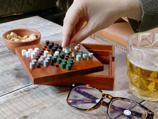 Една ръка движи фигура на дървен настолен комплект Игра Iron & Glory - Chinese Checkers, със закуски, чаша бира и очила на селска дървена маса - идеален подарък за любителите на игрите.-5