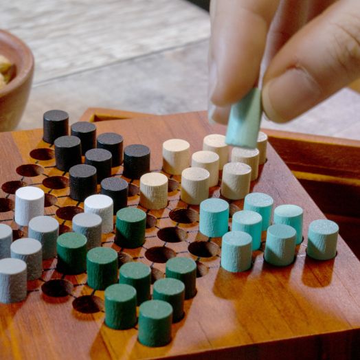 Една ръка поставя светлосин цилиндър върху Iron & Glory - Chinese Checkers дървен комплект, с редици от черни, бели, сини и зелени колчета - идеален за логически игри или като специален подарък.-4