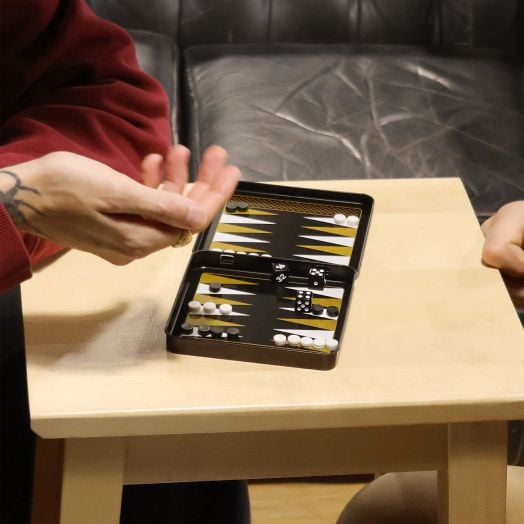 Двама души играят табла на малка дървена маса, използвайки Игра Iron & Glory - Магнитна табла. Едната ръка се готви да хвърли заровете. Компактната дъска с черни и бели фигури лежи до черен кожен диван.-5