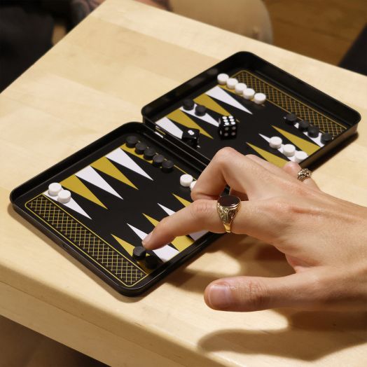 Човек премества фигура върху Игра Iron & Glory - Магнитна табла, компактна черна дъска за табла с бели и жълти шарки. Черните и белите фигури плюс заровете са разположени върху дървена маса - идеална за табла пътуване.-6