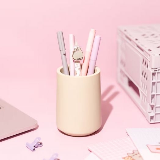 В чаша с кремав цвят са поставени сиви и розови химикалки, включително една изтриваема гел химикалка Grupo Erik Pusheen Moments с дизайн на сладка карикатурна котка, върху розово бюро с лаптоп, щипки за хартия и бял сандък-идеален подарък.-6