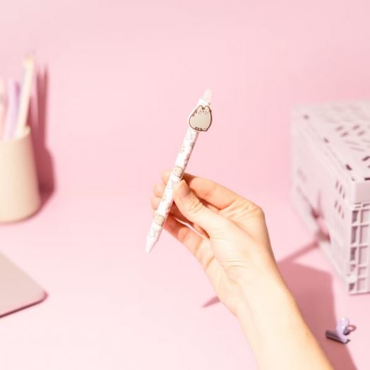 Една ръка държи Изтриваемата гел химикалка Grupo Erik Pusheen Moments, асортимент - със сладки анимационни мотиви на котки и талисман във формата на котка - на мек розов фон; идеален подарък за любителите на котки.-7