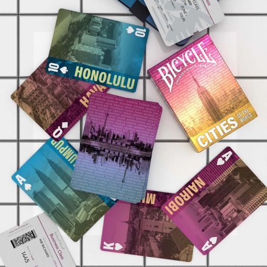 Карти за игра Bicycle Cities of the World с дизайни от градове като Хонолулу, Куала Лумпур, Найроби и Хавана са разположени върху бели плочки до бордна карта.-7