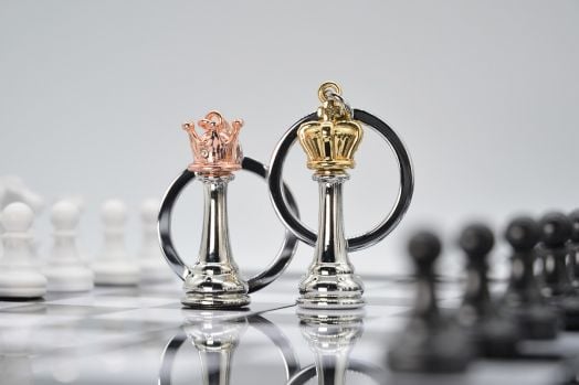 Два ключодържателя Metalmorphose - Queen Chess, с корони в розово злато и злато, стоят върху шахматна дъска - стилен подарък за фенове на шахмата. На заден план се виждат размазани фигури.-6