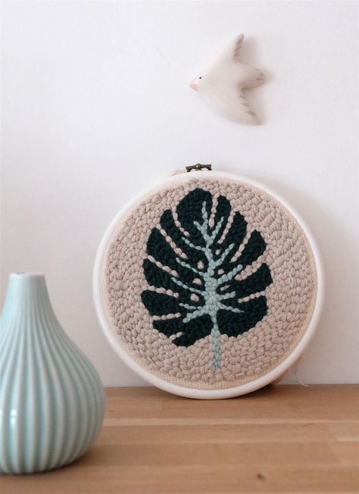Комплектът за пънч бродерия Graine Creative - Листо включва рамкирана, кръгла творба с перфорирана игла на тъмнозелено листо, разположено върху дърво до светлосиня ваза и бяла птица на мек фон.-4