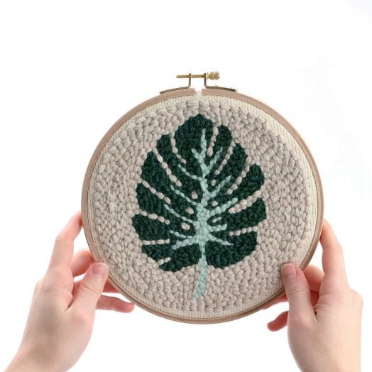 Два ръце държат комплект за пънч бродерия Graine Creative - Листо, показващ дизайн на зелено монстера листо върху кремав плат.-3