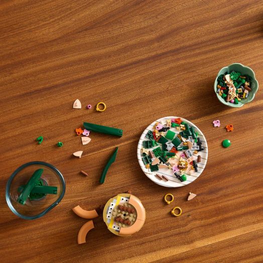 Частите от комплекта „LEGO Botanicals Миниорхидея“ в зелено, бежово и розово са подредени в малки купички върху дървена маса — вдъхновяващ подарък за възрастни, готов за творческо сглобяване.-5