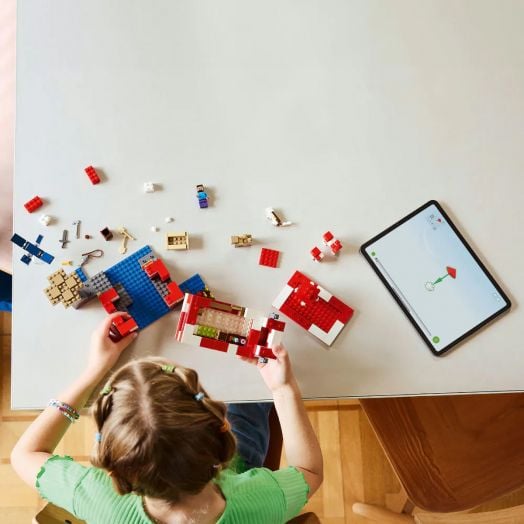 Дете с плитки седи на маса и строи с кубчета LEGO Minecraft „Къщата на Муухоморка“. Наоколо са разпръснати различни части, а на таблета се показват инструкциите за сглобяване на комплекта.-7