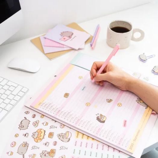 Човек пише на бяло бюро в пастелен цвят Планер-пад Grupo Erik Pusheen The Cat за 16 месеца (септември 2025 - декември 2026) с мотиви на карикатурна котка, заобиколен от клавиатура, мишка, химикалки, стикери, тетрадки и кафе.-3